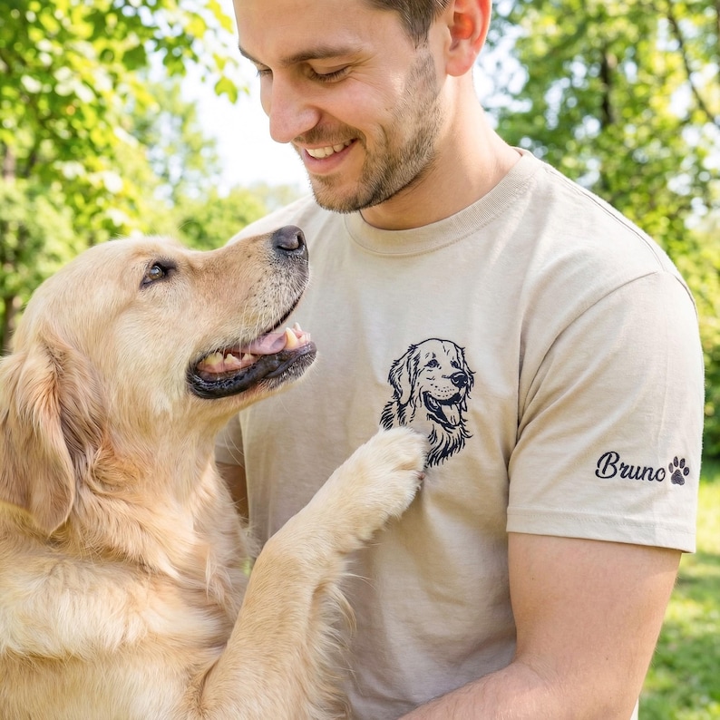 A sand-colored t-shirt laid on grass featuring a black thread embroidered Golden Retriever portrait on the left chest. The left sleeve is embroidered with the name “Bruno” and a small paw print detail.