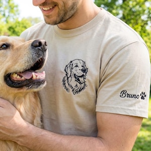 A sand-colored t-shirt laid on grass featuring a black thread embroidered Golden Retriever portrait on the left chest. The left sleeve is embroidered with the name “Bruno” and a small paw print detail.