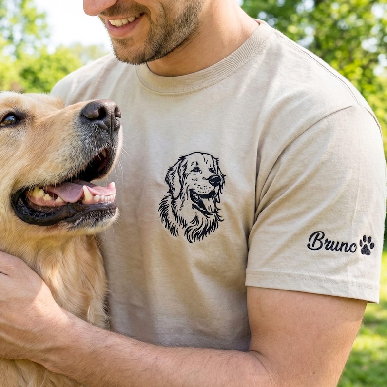 A sand-colored t-shirt laid on grass featuring a black thread embroidered Golden Retriever portrait on the left chest. The left sleeve is embroidered with the name “Bruno” and a small paw print detail.