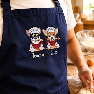 Puede incluir: Un delantal azul marino con un diseño bordado de dos perros pequeños con gorros de chef blancos y pañuelos rojos. Cada perro sostiene una cuchara de madera en la boca. Los nombres Jemma y Jax están bordados en blanco debajo de cada perro.