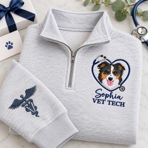 Puede incluir: Una sudadera gris claro con cremallera corta que presenta un diseño bordado de un perro dentro de un corazón de estetoscopio, con el texto Sophia VET TECH. La manga muestra un símbolo de caduceo bordado con una huella en el centro.