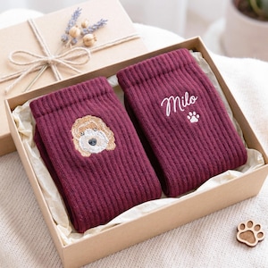 Könnte beinhalten: Ein Paar gerippte bordeauxrote Socken in einer Geschenkschachtel aus Karton. Eine Socke zeigt ein gesticktes goldbraunes Hundegesicht, während die andere den weiß gestickten Namen Milo über einem kleinen weißen Pfotenabdruck-Symbol trägt.