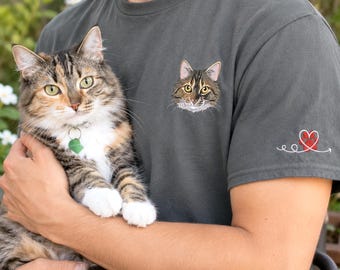 May include: A charcoal gray t-shirt featuring a small embroidered cat face on the left chest and a white line art graphic of a heart with a red paw print on the sleeve. The shirt is made of a solid fabric and has a crew neckline.