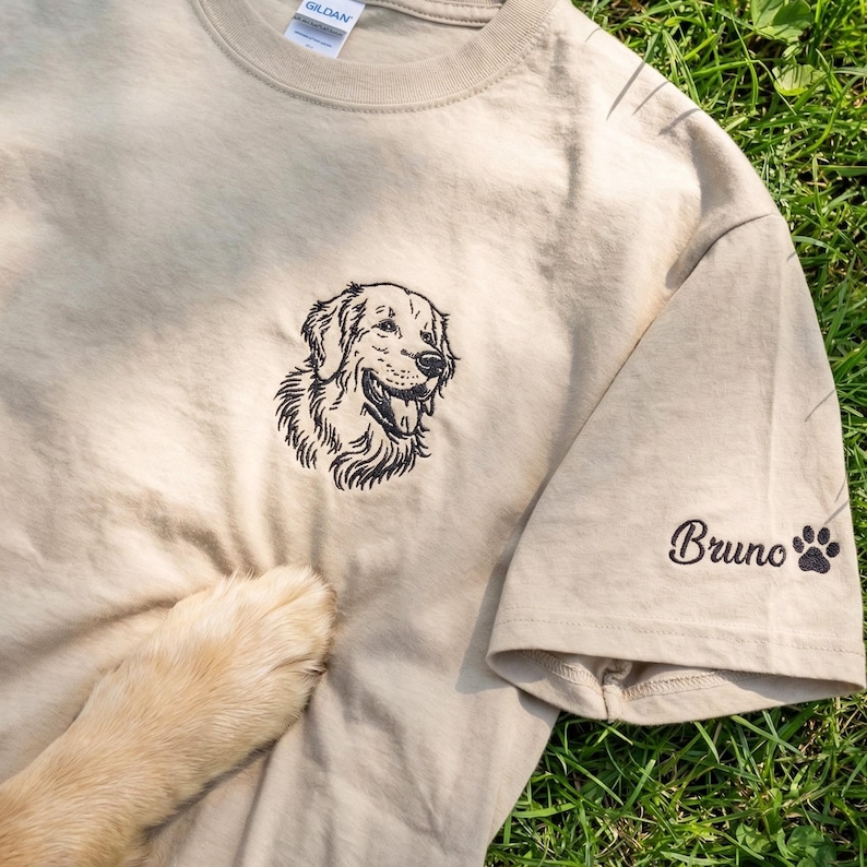 A sand-colored t-shirt laid on grass featuring a black thread embroidered Golden Retriever portrait on the left chest. The left sleeve is embroidered with the name “Bruno” and a small paw print detail.