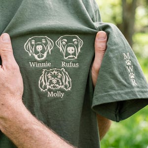 以下が含まれることがあります： 犬の顔と名前が刺繍されたグリーンのTシャツ。Winnie、Rufus、Molly。袖には足跡とイニシャルMWRがデザインされています。Tシャツは人が持っています。
