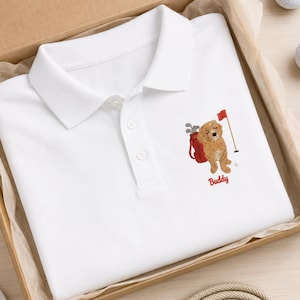 May include: A white polo shirt featuring an embroidered design of a golden-coloured dog standing next to a red golf bag with clubs and a red flag on a pole. The name Buddy is embroidered in red script lettering below the dog graphic.