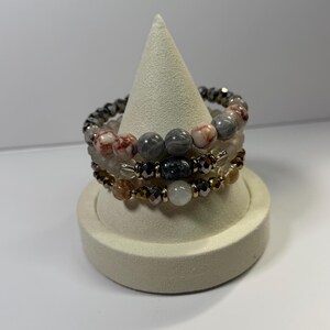 May include: Stack of three beaded bracelets in neutral tones, displayed on a white cone-shaped jewelry stand. The bracelets feature a variety of beads, including gray, white, and brown, with metallic accents. The bracelets are a mix of stone and crystal beads.