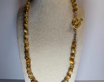 May include: A beaded necklace featuring golden-brown tiger eye stones and gold-colored accents. The necklace is displayed on a white jewelry bust and has a gold toggle clasp. The beads are approximately 0.3 inches in diameter.