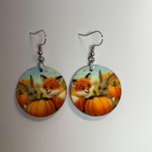 May include: A pair of round earrings featuring a fall theme. Each earring displays a smiling fox and a gray bunny next to an orange pumpkin. The background has a blue sky and autumn leaves.
