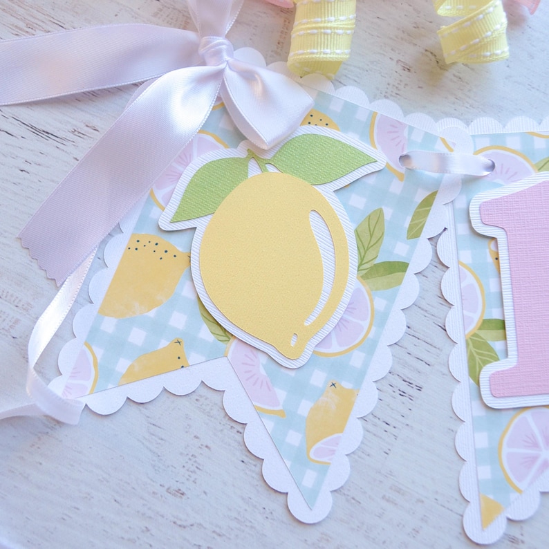 May include: A light blue and white gingham banner with a yellow lemon, green leaves, and a pink number one. The banner is decorated with a white scalloped edge.