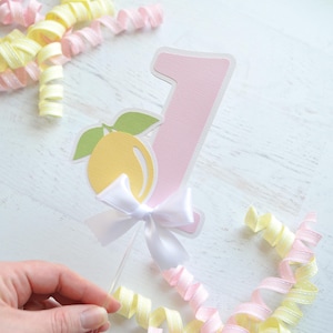 May include: A pink number one cake topper with a yellow lemon and green leaves, a white bow, and a clear plastic stick.