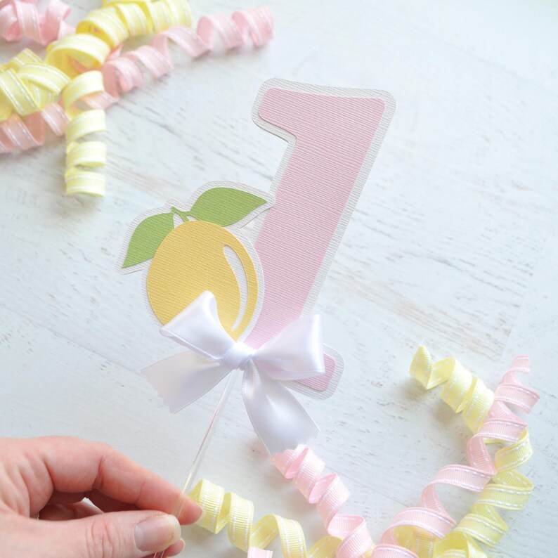 May include: A pink number one cake topper with a yellow lemon and green leaves, a white bow, and a clear plastic stick.