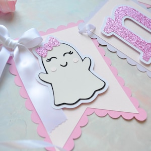 Pastel ghost first birthday high chair banner with pink and lavender scalloped backing, glitter pink letters spelling ONE, white satin bows, and smiling ghosts with pink glitter bows and rosy cheeks.