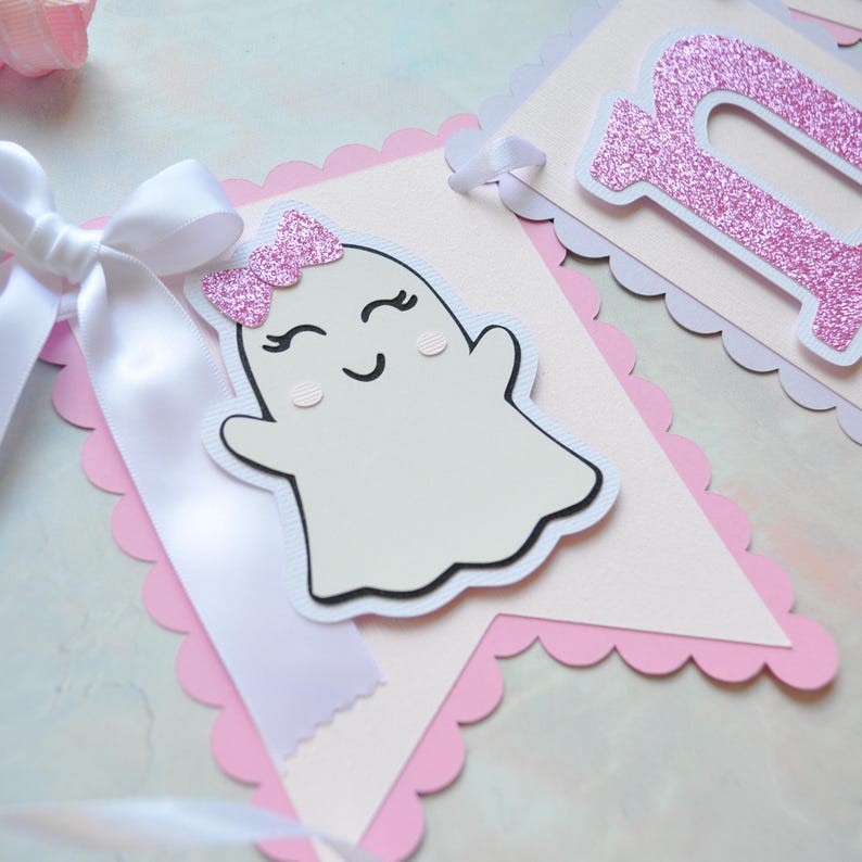 Pastel ghost first birthday high chair banner with pink and lavender scalloped backing, glitter pink letters spelling ONE, white satin bows, and smiling ghosts with pink glitter bows and rosy cheeks.