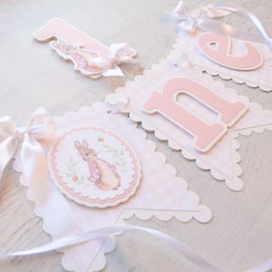 May include: A decorative banner with a Peter Rabbit theme. The banner features pennants with pink gingham patterns, scalloped edges, and white satin ribbons. The banner includes the number "1" and the letter "n" in pink, along with a circular image of Peter Rabbit.