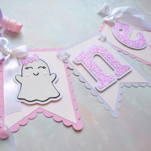 Pastel ghost first birthday high chair banner with pink and lavender scalloped backing, glitter pink letters spelling ONE, white satin bows, and smiling ghosts with pink glitter bows and rosy cheeks.