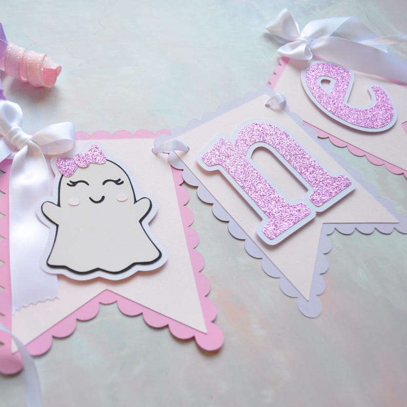 Pastel ghost first birthday high chair banner with pink and lavender scalloped backing, glitter pink letters spelling ONE, white satin bows, and smiling ghosts with pink glitter bows and rosy cheeks.