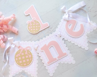 May include: A decorative banner set featuring the word one in light pink letters on white scalloped pennants with a light pink gingham pattern. The set includes a separate number one cutout with a gold waffle-textured circle and pink bow.