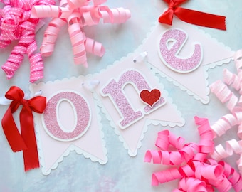 May include: Pink and white banner with the word "one" in pink glitter letters. A red heart is on the "n". The banner is decorated with pink and red curled ribbon.