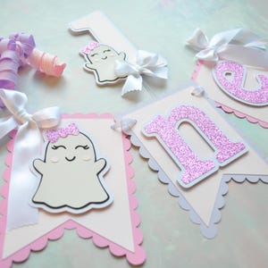 Pastel ghost first birthday high chair banner with pink and lavender scalloped backing, glitter pink letters spelling ONE, white satin bows, and smiling ghosts with pink glitter bows and rosy cheeks.

White and pink number one cake topper with cute smiling ghost wearing a pink glitter bow, accented with a white satin bow, perfect for a pastel Halloween first birthday cake.