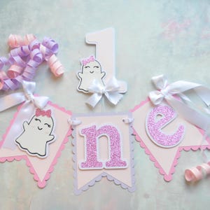 Pastel ghost first birthday high chair banner with pink and lavender scalloped backing, glitter pink letters spelling ONE, white satin bows, and smiling ghosts with pink glitter bows and rosy cheeks.

White and pink number one cake topper with cute smiling ghost wearing a pink glitter bow, accented with a white satin bow, perfect for a pastel Halloween first birthday cake.