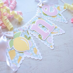 May include: A light blue and yellow banner with the word "one" in pink letters. The banner has a scalloped edge and a lemon pattern.