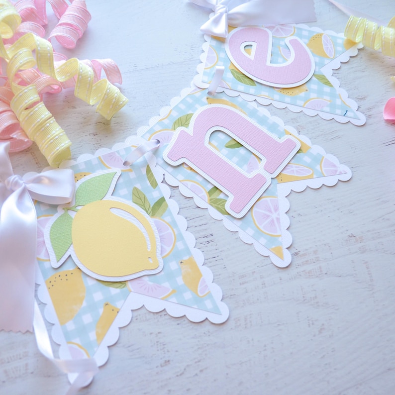 May include: A light blue and yellow banner with the word "one" in pink letters. The banner has a scalloped edge and a lemon pattern.