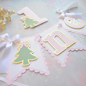 May include: A festive banner with pink, white, and gold accents. The banner features the number "1", the letter "n", and a letter "d", along with a green Christmas tree with a pink bow. White ribbon is attached to the banner.