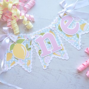 May include: A light blue and white gingham banner with pink letters spelling "one" and a lemon print. The banner is decorated with white bows and yellow and pink curling ribbon.