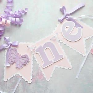 May include: A pink and purple banner with the word "one" spelled out in purple letters. Each letter is on a separate pink pennant with a scalloped edge. The banner is decorated with a purple butterfly on the first pennant.
