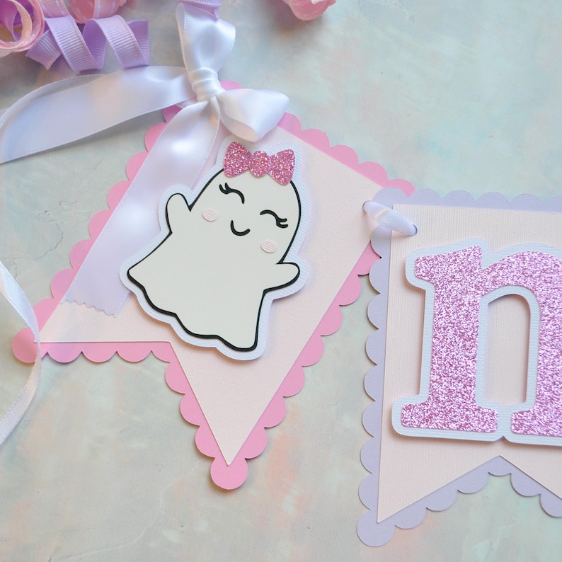 Pastel ghost first birthday high chair banner with pink and lavender scalloped backing, glitter pink letters spelling ONE, white satin bows, and smiling ghosts with pink glitter bows and rosy cheeks.