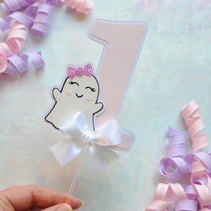 White and pink number one cake topper with cute smiling ghost wearing a pink glitter bow, accented with a white satin bow, perfect for a pastel Halloween first birthday cake.
