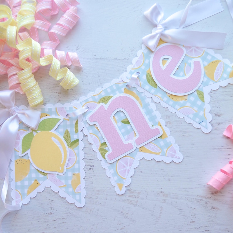 May include: A light blue and white gingham banner with pink letters spelling "one" and a lemon design. The banner has scalloped edges and is decorated with white ribbon bows.
