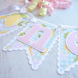 May include: A light blue and white banner with a lemon pattern and pink felt letters spelling "one". The banner has a scalloped edge and is strung with white ribbon.
