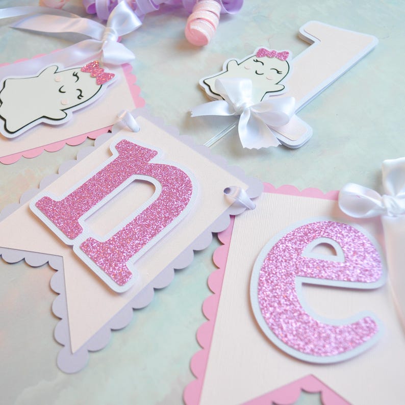 Pastel ghost first birthday high chair banner with pink and lavender scalloped backing, glitter pink letters spelling ONE, white satin bows, and smiling ghosts with pink glitter bows and rosy cheeks.

White and pink number one cake topper with cute smiling ghost wearing a pink glitter bow, accented with a white satin bow, perfect for a pastel Halloween first birthday cake.