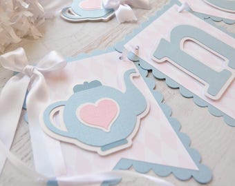 May include: A pastel blue and pink banner featuring a teapot and heart motif. The banner has a scalloped edge and is adorned with white ribbon. This is part of a party decoration set.