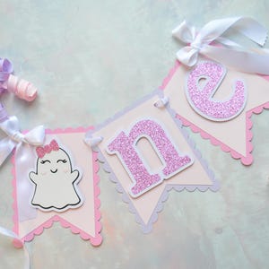 Pastel ghost first birthday high chair banner with pink and lavender scalloped backing, glitter pink letters spelling ONE, white satin bows, and smiling ghosts with pink glitter bows and rosy cheeks.