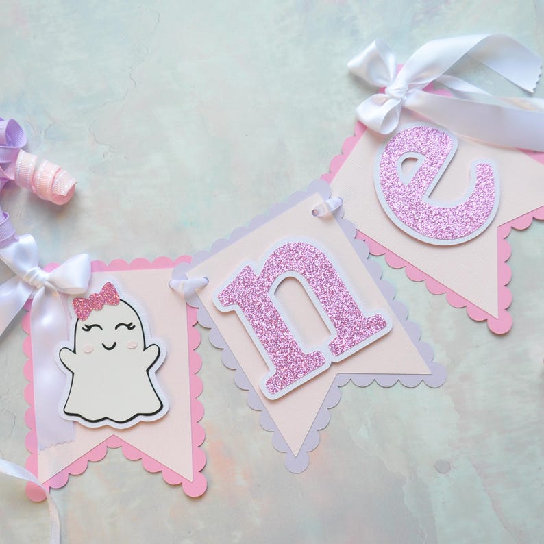 Pastel ghost first birthday high chair banner with pink and lavender scalloped backing, glitter pink letters spelling ONE, white satin bows, and smiling ghosts with pink glitter bows and rosy cheeks.