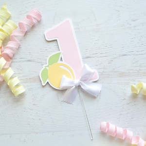 May include: Pink paper number one with a yellow lemon and a white bow. The number one is attached to a white stick.