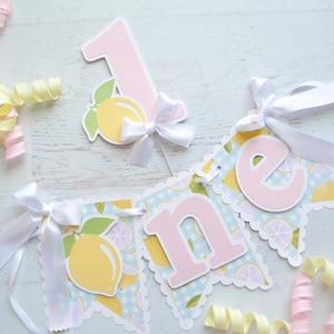 May include: A pink and yellow lemon-themed banner with the word "one" in pink letters. The banner is decorated with white ribbon and lemons on a blue and white gingham background.