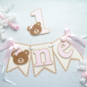 May include: A pink and brown 'one' banner with a brown bear head with a pink bow on each side of the word 'one'. The banner is decorated with scalloped edges and pink ribbon bows. A separate pink and brown '1' with a brown bear head with a pink bow is also included.