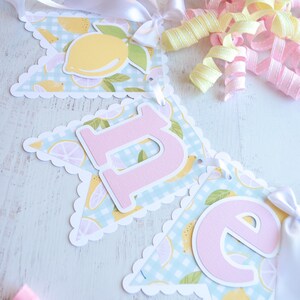 May include: A light blue and white banner with a lemon print and pink letters spelling "Lime". The banner has a scalloped edge and is strung with white ribbon.