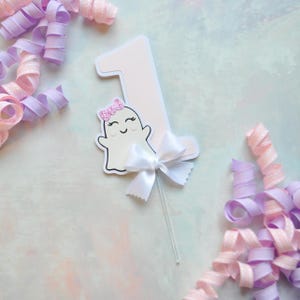 White and pink number one cake topper with cute smiling ghost wearing a pink glitter bow, accented with a white satin bow, perfect for a pastel Halloween first birthday cake.