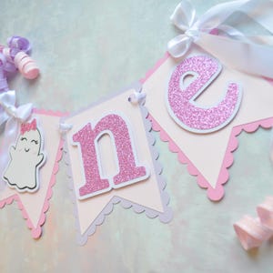 Pastel ghost first birthday high chair banner with pink and lavender scalloped backing, glitter pink letters spelling ONE, white satin bows, and smiling ghosts with pink glitter bows and rosy cheeks.