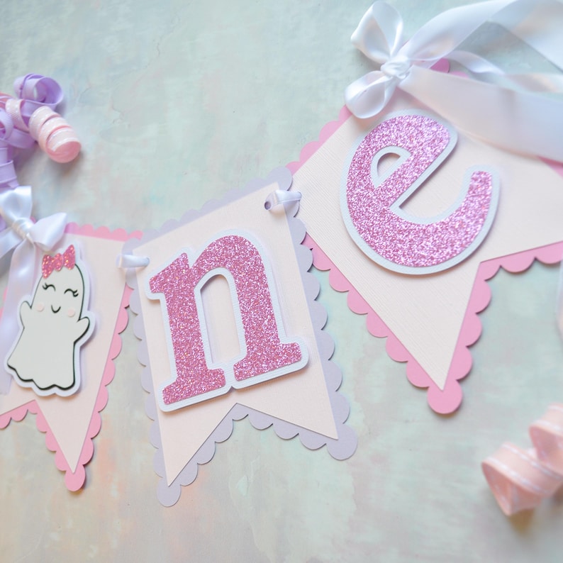 Pastel ghost first birthday high chair banner with pink and lavender scalloped backing, glitter pink letters spelling ONE, white satin bows, and smiling ghosts with pink glitter bows and rosy cheeks.