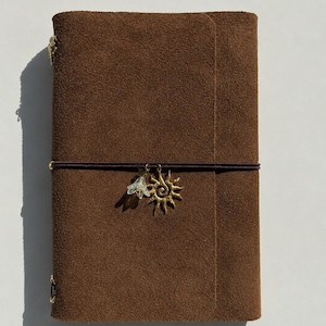 May include: A rectangular brown suede leather journal cover featuring a dark brown elastic band wrapped horizontally around the center. Two metal charms are attached to the elastic: a gold-toned sunburst charm and a small, translucent white flower.