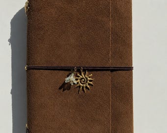 May include: A rectangular brown suede leather journal cover featuring a dark brown elastic band wrapped horizontally around the center. Two metal charms are attached to the elastic: a gold-toned sunburst charm and a small, translucent white flower.