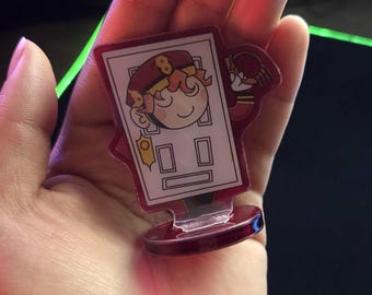 Könnte beinhalten: Ein klarer Acryl-Anhänger mit einer Cartoonfigur mit rotem Hut und Uniform, die in einer Tür steht. Der Anhänger hat einen roten Rand und eine rote Basis und wird in einer Hand gehalten.