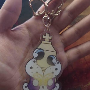 May include: A gold-coloured keyring featuring a cartoon chess king design. The king is yellow with a purple and white crown and large, expressive eyes. The keyring has a gold-coloured clasp.