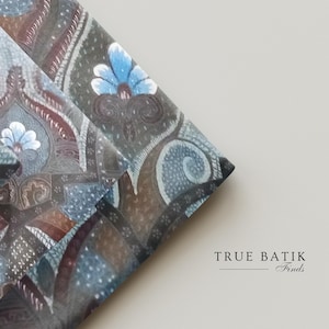 Op de afbeelding: Close-up van gevouwen stof met een gedetailleerd batikpatroon. Het ontwerp bevat bloemen- en paisley-motieven in tinten bruin, blauw en wit. De tekst "TRUE BATIK Finds" is rechtsonder zichtbaar.